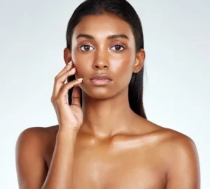 A woman with long hair poses against a plain background, touching her face with one hand and gazing directly at the camera.