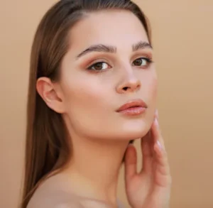 A woman with straight brown hair poses against a beige background, touching her chin with one hand, displaying a neutral expression.