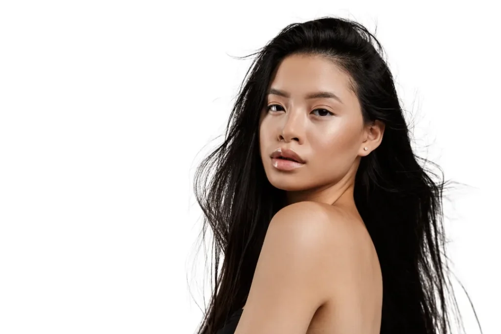 A person with long dark hair and bare shoulders looks over their shoulder against a white background.